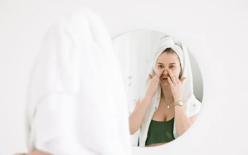 A woman in a towel performing her skincare routine in front of a bathroom mirror.