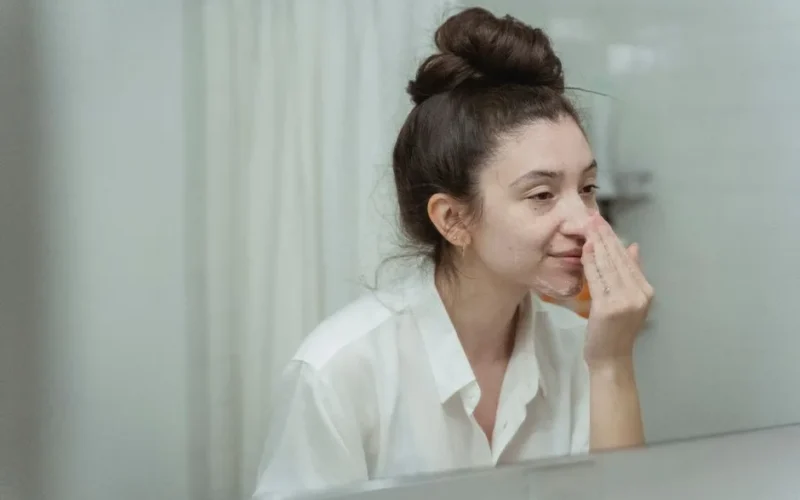 Woman applying face cream in bathroom, focusing on skincare routine with natural reflection in mirror.