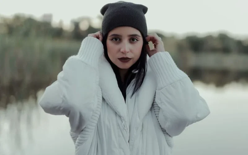A woman in a white coat and beanie poses by a serene lakeside, embodying winter fashion.