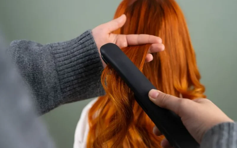 Hands styling long red hair using a flat iron indoors. Haircare and beauty concept.