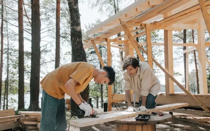 Two carpenters working outdoors constructing a wooden frame in a forest setting.