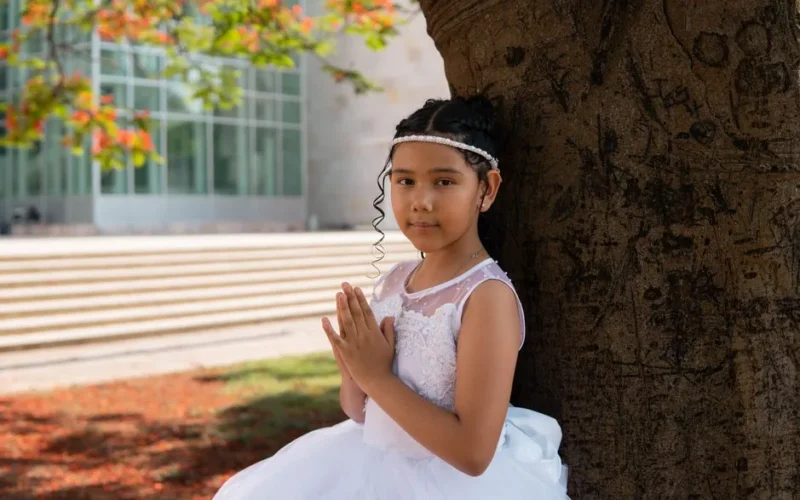 A young girl in a white dress poses elegantly against a tree for her First Communion portrait.