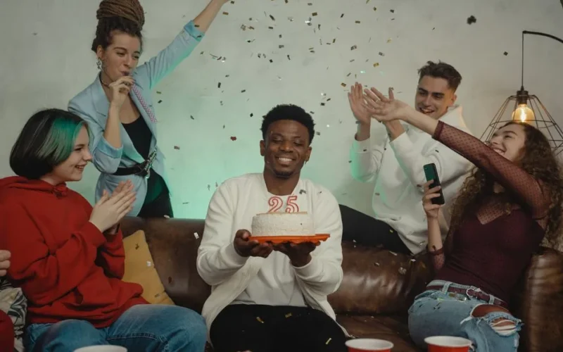 A diverse group of friends celebrates a 25th birthday indoors with cake and confetti.