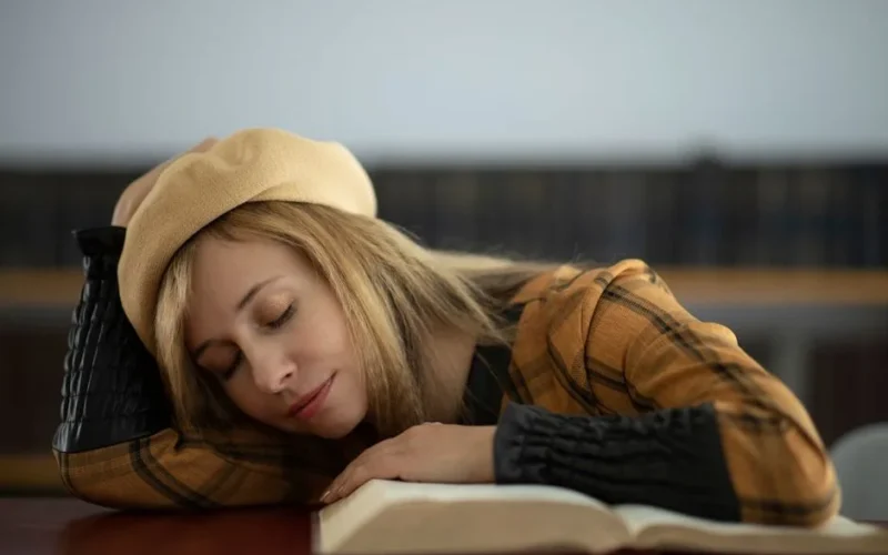 A young woman in a library naps on an open book with eyes closed, wearing a beret hat.