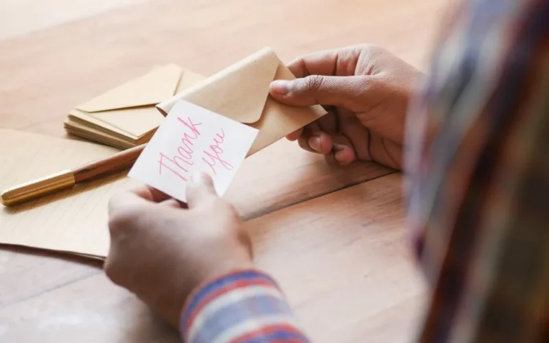 Close-up of hands holding a thank you note in an envelope on a table.