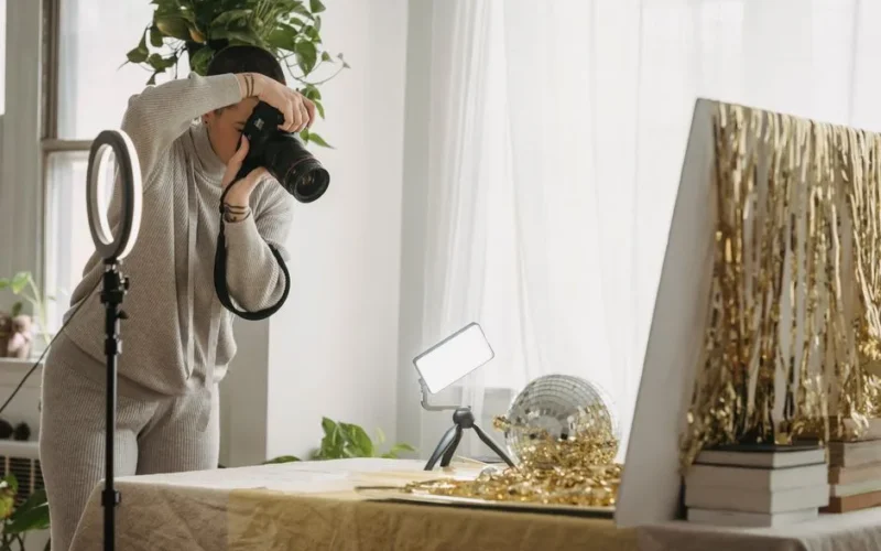 Female photographer with short hair standing at table with shimmering tinsel and party ball near luminous lamp on tripod