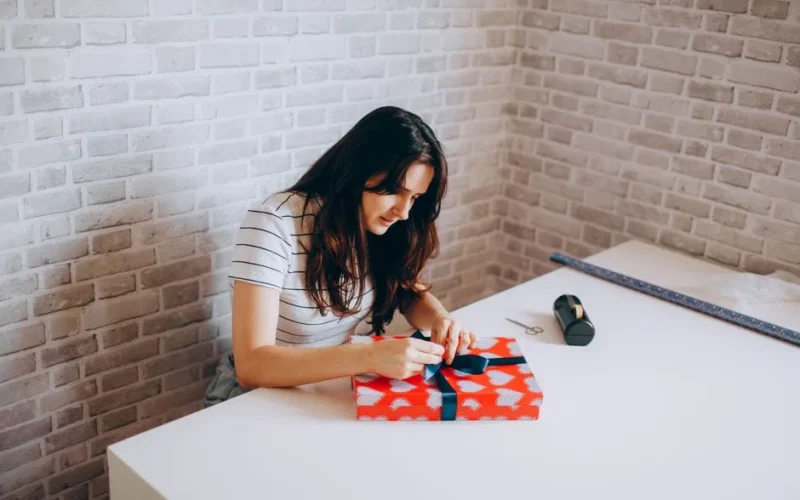 Woman gift wrapping at table in cozy room with brick walls, showcasing creativity and leisure.