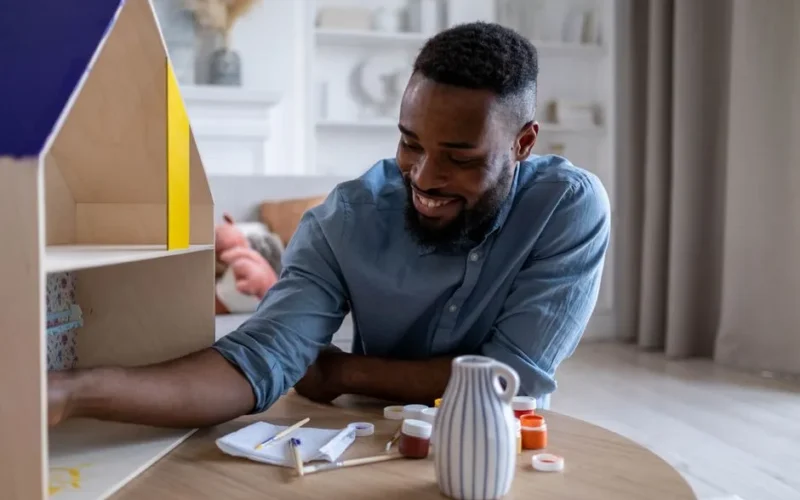 A joyful man enjoys crafting a dollhouse indoors, expressing creativity and happiness.