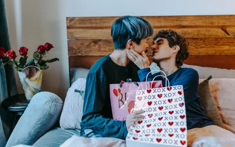 A loving couple exchanges Valentine's gifts on a cozy bed adorned with roses and heart-themed gift bags.