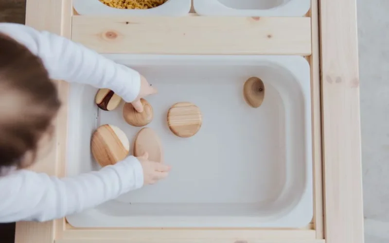 From above view of unrecognizable little child playing with wooden round stones in white plastic container at wooden table at home