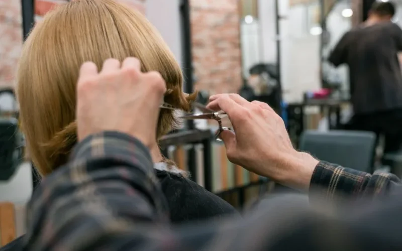 Hairdresser cutting woman's blonde hair in salon with scissors and mirror.