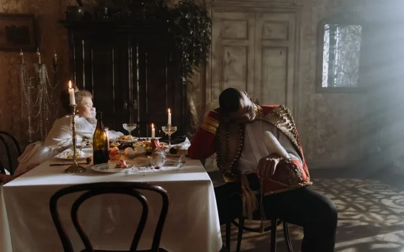 Two adults in period costumes asleep after a party at an elegant dining room table.
