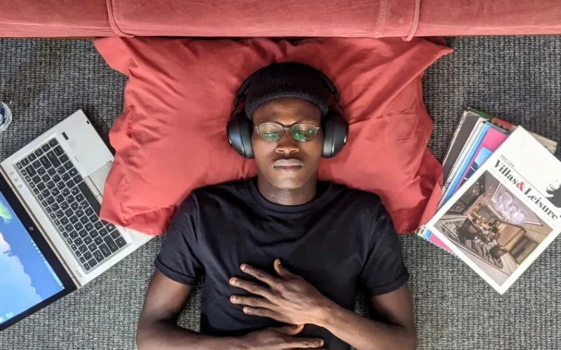A young man wearing headphones, relaxing with eyes closed, laptop and magazines by his side.