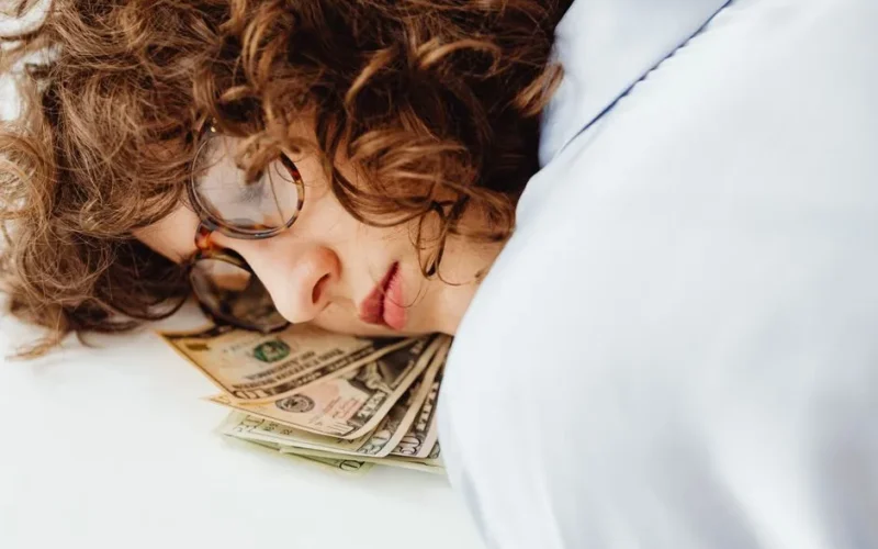 Close-up of a woman with curly hair and glasses resting her head on US dollar bills, symbolizing dreams of wealth.