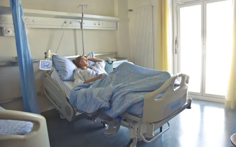 A woman lying in a hospital bed in a bright, softly lit room, appearing to rest or recover.