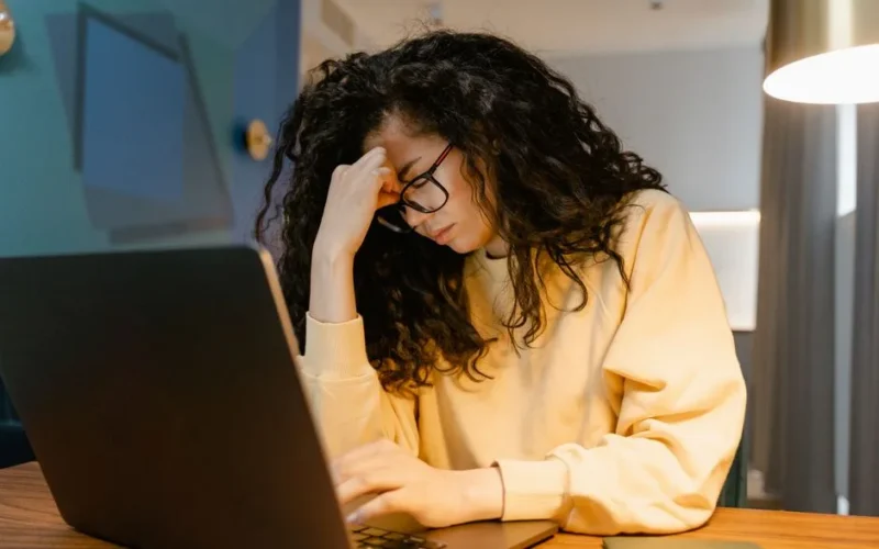 A tired woman with curly hair and glasses experiencing stress while working on a laptop.