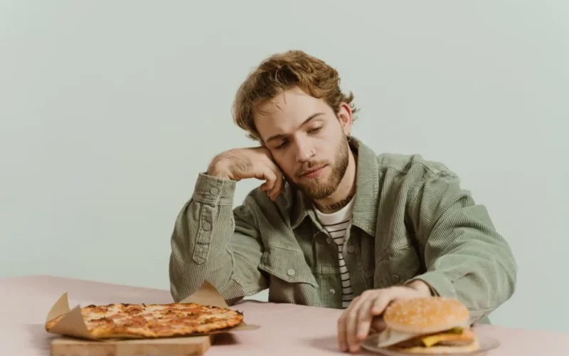 A man sits thoughtfully at a table with a pizza and burger in a minimalist indoor setting.
