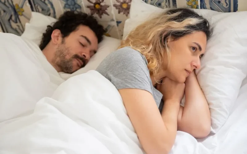 A woman lies awake next to her sleeping partner in a cozy bedroom setting, contemplating.