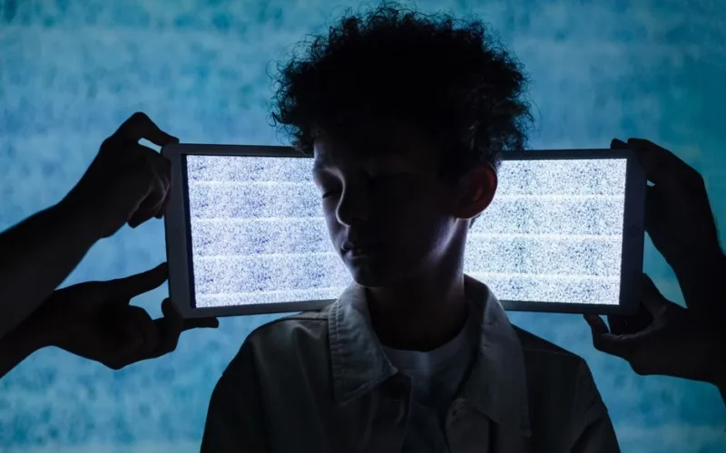 A teenager stands with eyes closed between two tablets showing digital noise, symbolizing information overload.
