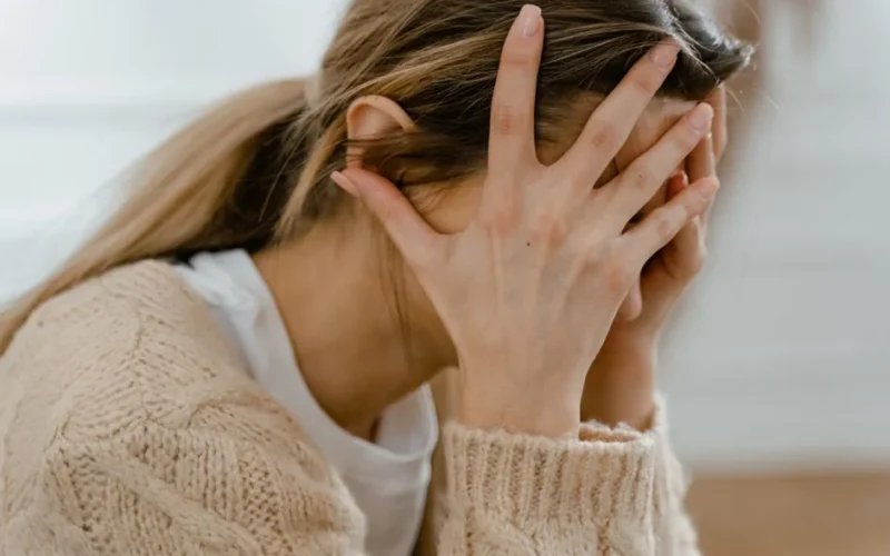 Woman sitting indoors with face covered by hands, expressing stress and frustration.