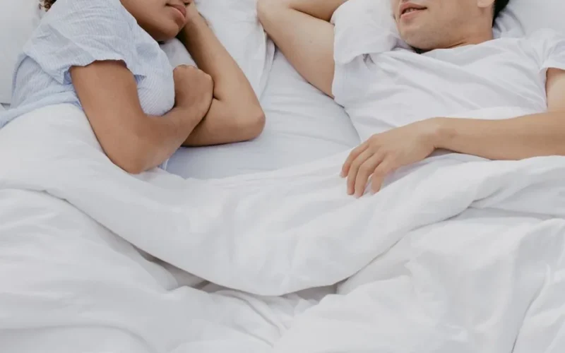 Two adults comfortably lying in bed together, enjoying a peaceful moment under white sheets.