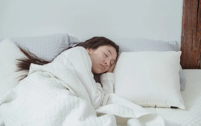 From above of young calm female with long dark wavy hair in white nightwear sleeping on soft pillow in bed under blanket in morning at home