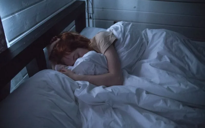 A woman sleeping peacefully in a cozy bedroom, enveloped by soft white sheets, under the gentle glow of night lighting.
