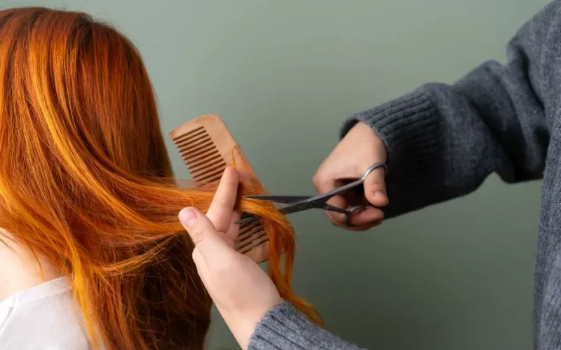 Close-up of red hair being trimmed with scissors and comb on green background.