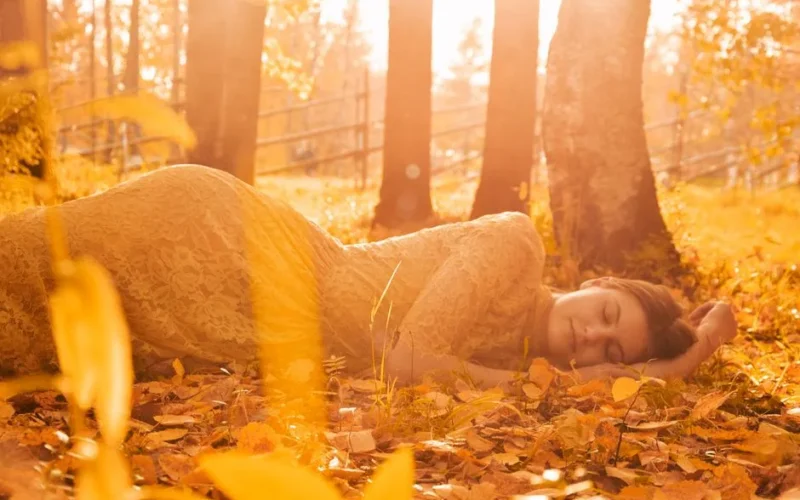 A woman peacefully lying on autumn leaves in a forest, basking in golden sunlight.