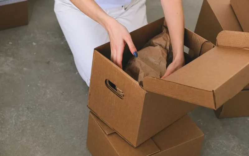 From above of crop anonymous relocating female packing wrapped items into carton box while preparing for moving to new apartment
