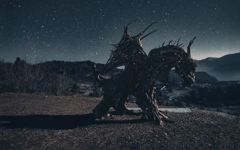 A mystical wooden sculpture resembling a creature under a star-filled sky in a mountainous landscape.