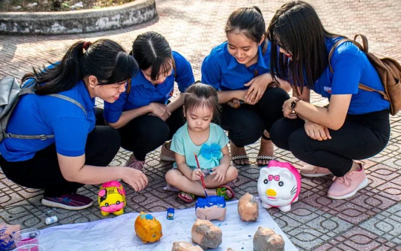 A child and women painting piggy banks together outdoors, enjoying a creative activity.