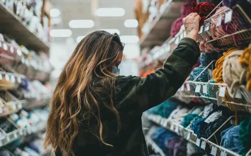 A woman wearing a mask browsing colorful yarn in a craft store aisle.