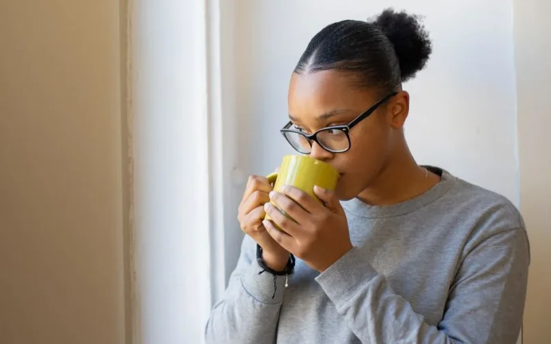A woman in glasses calmly savoring a hot drink in a cozy indoor setting by a window.