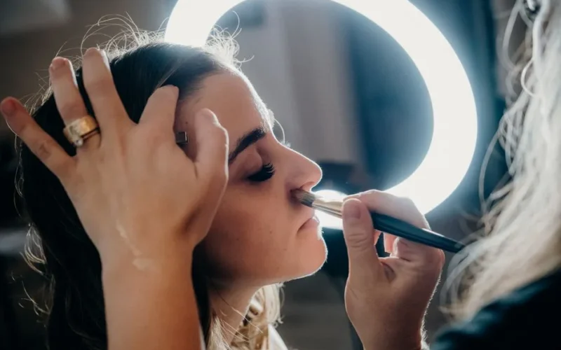Close-up of a woman receiving makeup under a ring light by a professional artist.