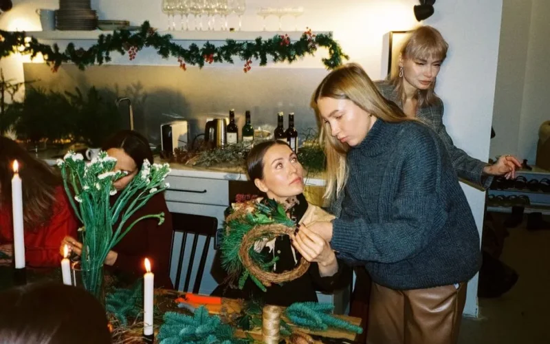 Women gather around to prepare handmade Christmas wreaths in a cozy indoor setting.