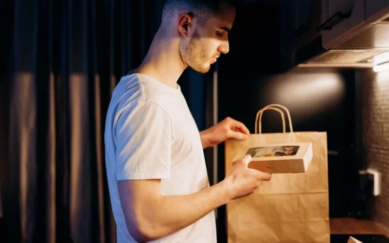 A man unpacks a food delivery from a paper bag in a warmly lit kitchen setting.