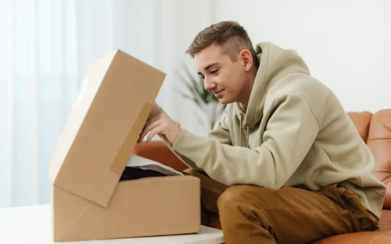 A young man happily unpacks a cardboard box while sitting on a couch indoors.