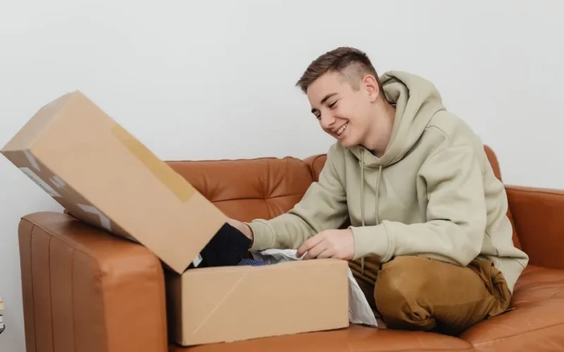 Smiling young man unboxing a package while sitting on a brown leather couch indoors.