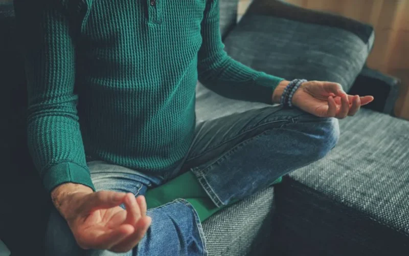 Close-up of a person meditating at home, cross-legged on a couch, emphasizing relaxation and mindfulness.