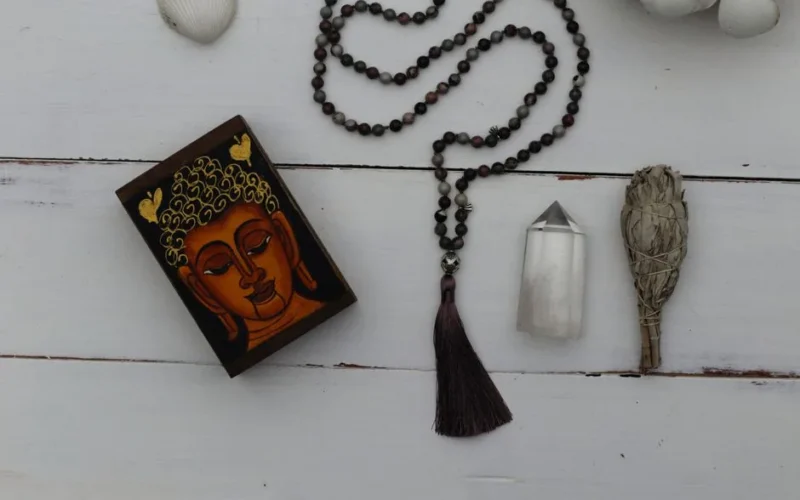 Flat lay of spiritual items including Buddha box, prayer beads, crystal, and sage bundle.