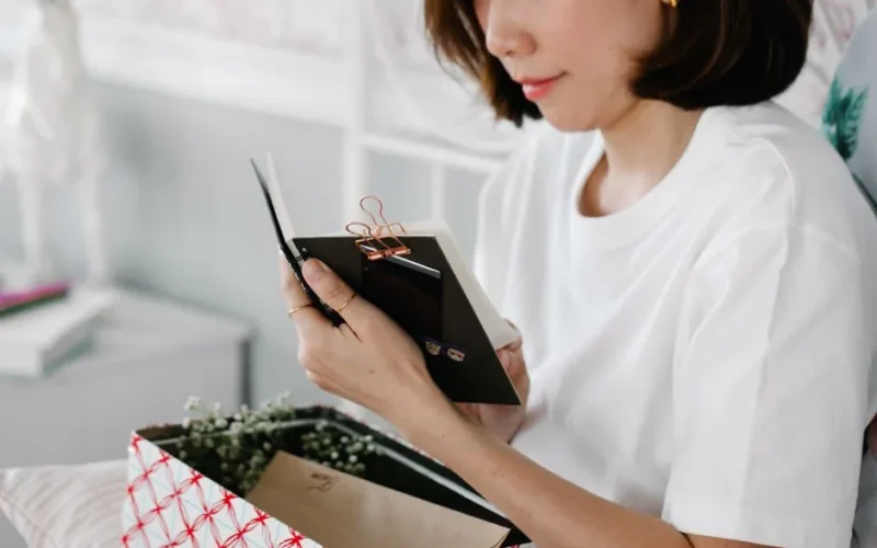 A woman in a white t-shirt opens a gift box and reads a notepad while relaxing indoors.