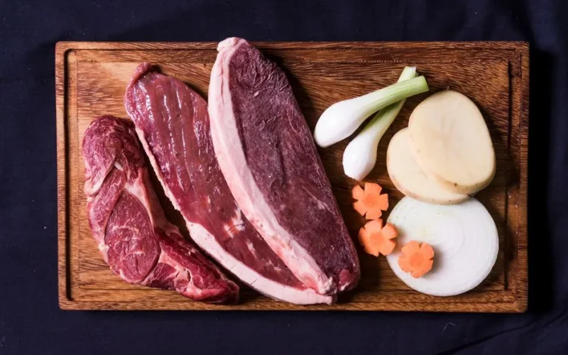 Top view of raw beef slices with root vegetables and spring onions arranged on a wooden board.