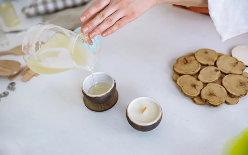 Close-up of hand pouring candle wax into rustic container, crafting essentials nearby.