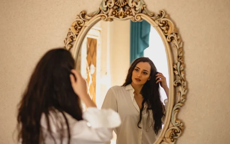 Elegant woman with long brunette hair gazing at her reflection in an ornate mirror, wearing white shirt indoors.