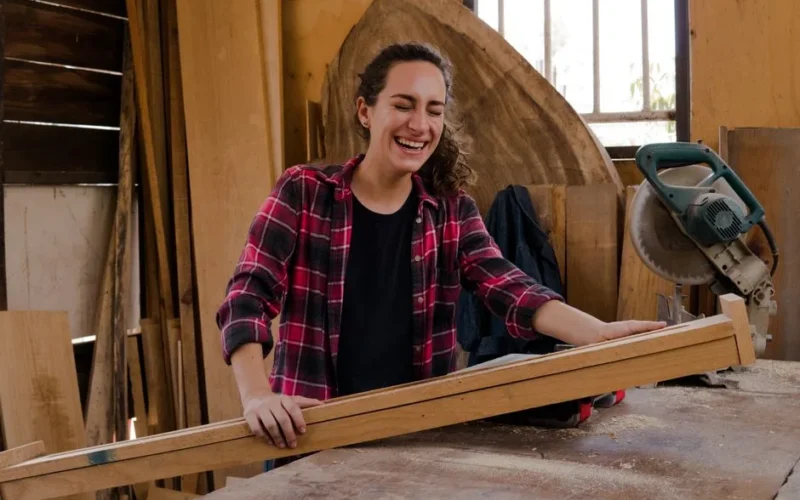 A happy woman in a plaid shirt working with wood in an indoor carpentry workshop.
