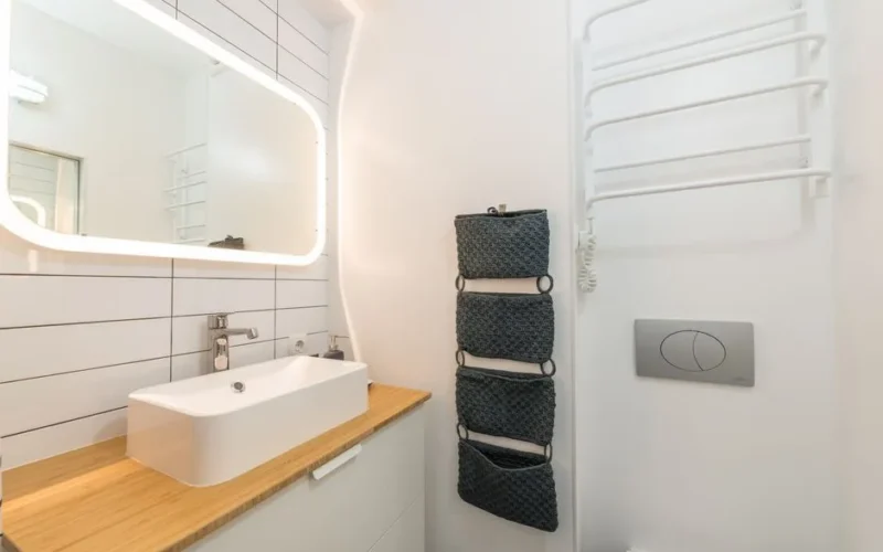 Interior of small modern bathroom with ceramic sink placed on white cabinet with wooden top under illuminated mirror