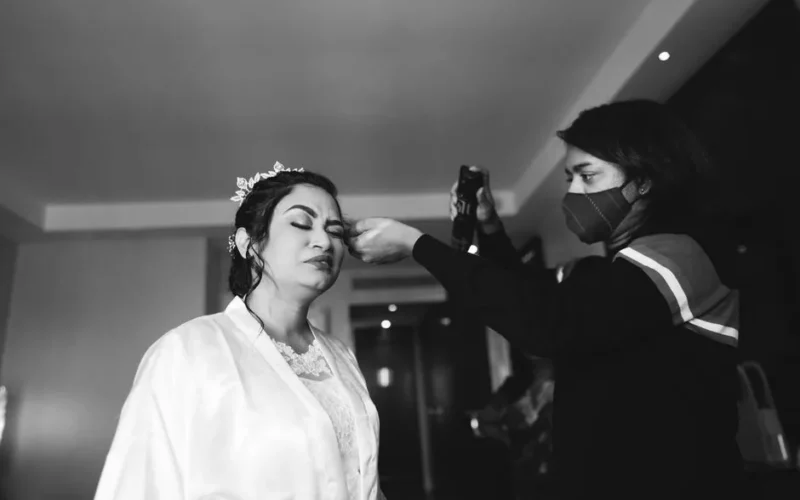 Bride getting makeup done indoors by a stylist wearing a mask.