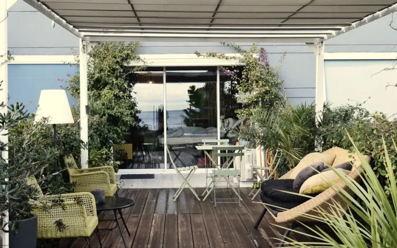 Comfortable stylish rattan armchairs with tables placed on wooden veranda of modern residential villa with lush exotic plants on sunny day