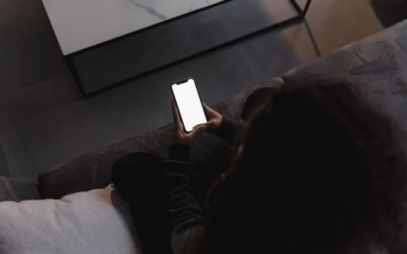 A person holding a smartphone with a blank screen, seated indoors on a couch.